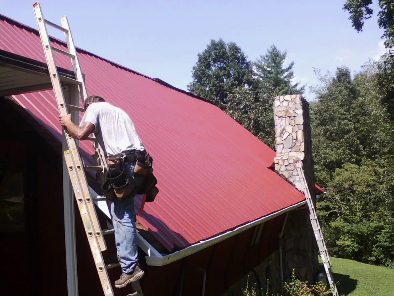 Roofer on a ladder beside a red metal roof and stone chimney — Soffit & Fascia Repair by Roofing Brothers in South Hackensack